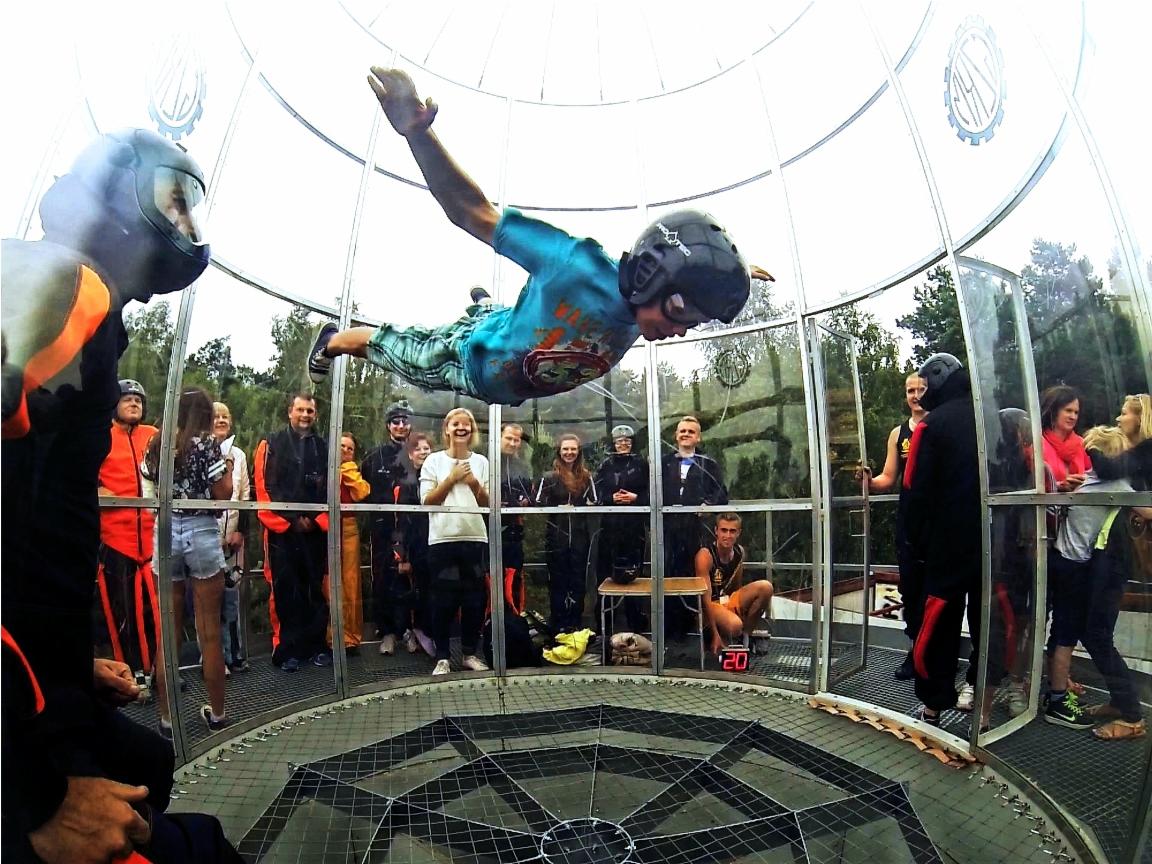 Mężczyzna w kasku i kombinezonie unosi się w tunelu aerodynamicznym, obserwuje go grupa osób za szklaną ścianą.