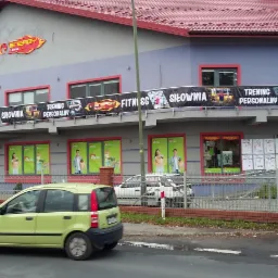 Banery reklamowe siłowni 'Energy Fitness Club' na budynku z czerwonym dachem. Żółty samochód na pierwszym planie, osoba przechodząca obok.
