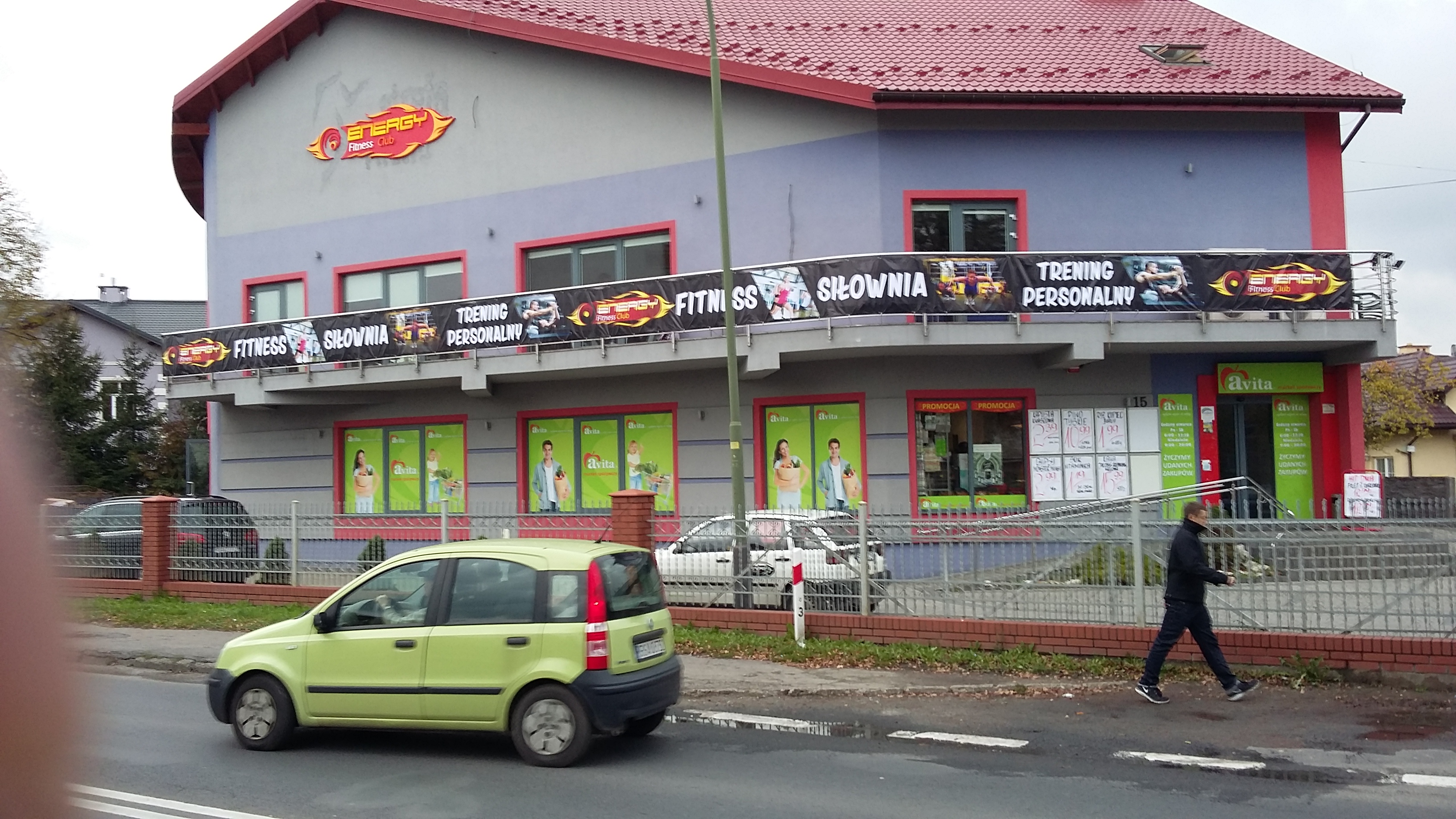 Banery reklamowe siłowni 'Energy Fitness Club' na budynku z czerwonym dachem. Żółty samochód na pierwszym planie, osoba przechodząca obok.