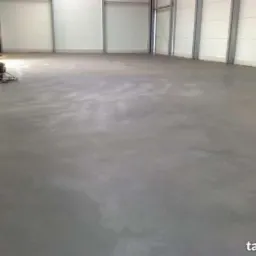 Wnętrze hali z szarą, gładką posadzką w trakcie polerowania. Widoczna maszyna do polerowania podłóg i metalowe słupy konstrukcyjne.