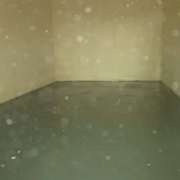 Świeżo wylana, ciemna posadzka w pustym pomieszczeniu o beżowych ścianach, z widocznym fragmentem węża po lewej stronie. Nierówności na powierzchni posadzki.