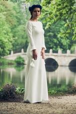 Elegancka kobieta w długiej, białej sukni z długimi rękawami i delikatnym wiankiem na głowie pozuje na tle malowniczego parku z jeziorem i kamiennym mostem.