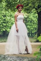 Modelka w białej, tiulowej sukni z gorsetem i asymetrycznym dołem, na tle zielonego parku, z wiankiem z czerwonych kwiatów na głowie.
