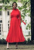 Modelka w długiej, czerwonej, plisowanej sukience bez rękawów z wiązaniem przy szyi, pozująca na tle klasycystycznego budynku i zieleni.