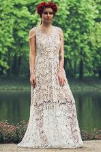 Kobieta w długiej, białej, koronkowej sukience z delikatnymi zdobieniami na ramionach, pozująca na tle zieleni i wody.