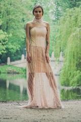 Modelka w długiej, zwiewnej, beżowej sukience z półprzezroczystego materiału, pozująca na tle parku z jeziorem i kamiennym mostkiem.
