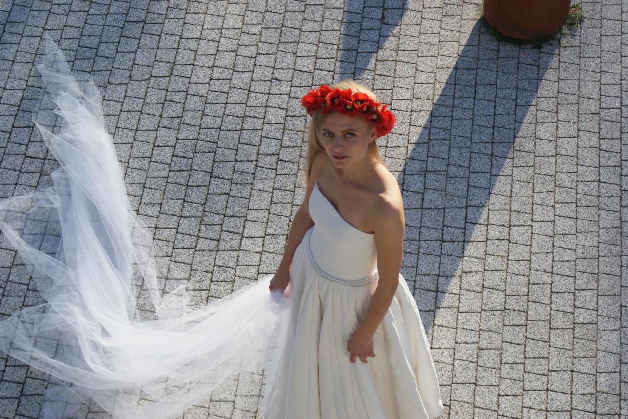 Panna młoda w białej sukni z jednym ramieniem i wiankiem z czerwonych maków, spoglądająca w górę, z długim, zwiewnym welonem na brukowanym dziedzińcu.