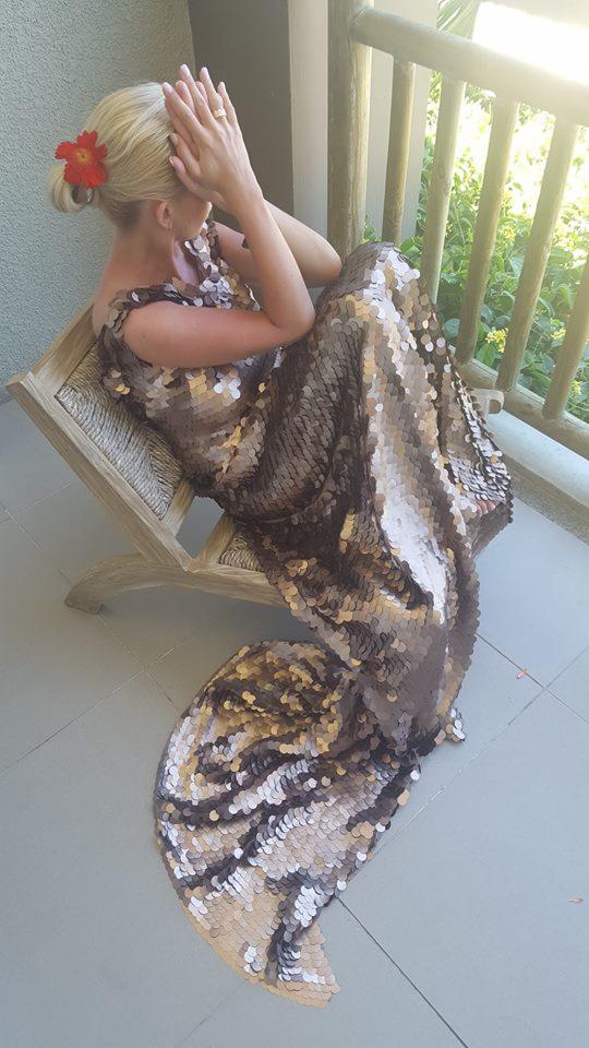 Kobieta siedząca na krześle na balkonie, ubrana w długą, cekinową suknię z trenem w odcieniach brązu i beżu, z czerwoną ozdobą we włosach.