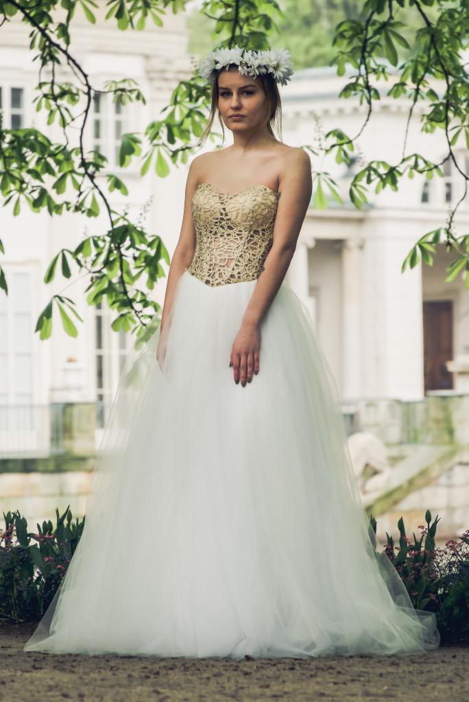 Młoda kobieta w białej sukni balowej z koronkowym, złotym gorsetem i wiankiem z białych kwiatów na głowie, pozująca na tle parku i klasycystycznego budynku.