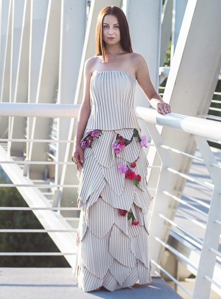 Kobieta w sukni bez ramiączek w paski z dekoracjami z różowych i czerwonych kwiatów, opierająca się o białą metalową konstrukcję na zewnątrz.