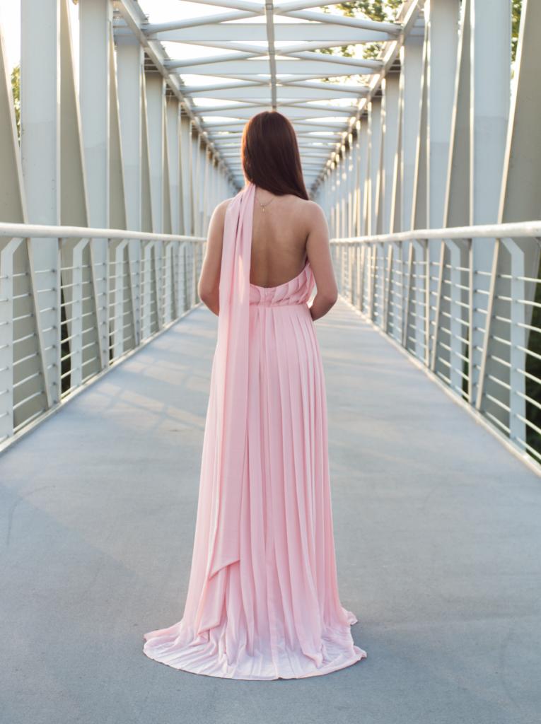Kobieta w długiej, plisowanej, pudrowo-różowej sukni z odkrytymi plecami, stojąca na metalowym moście, widok z tyłu.