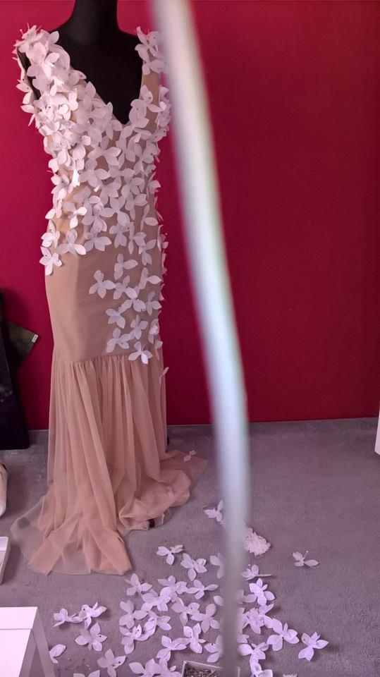 Suknia wieczorowa w kolorze beżowym, zdobiona białymi kwiatami, prezentowana na manekinie na tle czerwonej ściany, z dodatkowymi kwiatami leżącymi na podłodze.