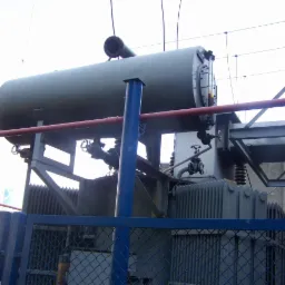 Duży transformator elektryczny w metalowej obudowie, zabezpieczony niebieskim ogrodzeniem z siatki. Widoczne elementy instalacji i rurociągi.