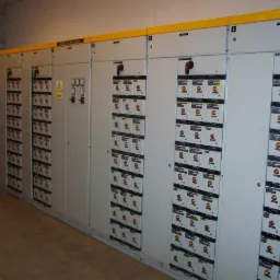 Rozbudowana, przemysłowa rozdzielnia elektryczna z licznymi zabezpieczeniami i wyłącznikami, oznaczona żółtymi elementami, umieszczona w pomieszczeniu technicznym.