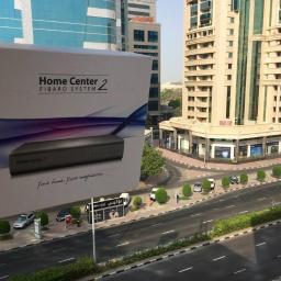 Sarko Krzysztof Okrasa - Pudełko z urządzeniem Home Center 2 FIBARO SYSTEM prezentowane na tle miejskiego krajobrazu z wysokimi budynkami i ulicą.