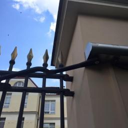 Sarko Krzysztof Okrasa - Zewnętrzny reflektor LED zamontowany na elewacji budynku, widoczny przez ozdobne, metalowe ogrodzenie z pozłacanymi grotami.