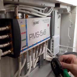 Sarko Krzysztof Okrasa - Instalacja multiswitcha Globo PMS-5x8 w skrzynce rozdzielczej, widoczne kable koncentryczne i narzędzie zaciskowe w ręce instalatora.