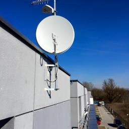 Sarko Krzysztof Okrasa - Antena satelitarna i antena kierunkowa zamontowane na szczycie budynku z szarej elewacji, widok z boku, na tle błękitnego nieba.