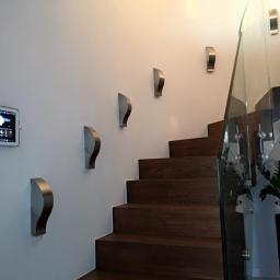Sarko Krzysztof Okrasa - Panel sterowania alarmem domowym, zamontowany na ścianie obok schodów z drewnianymi stopniami i szklaną balustradą, z dodatkowym oświetleniem w postaci metalowych kinkietów.