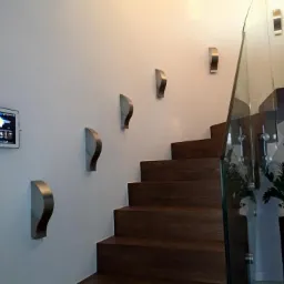 Panel sterowania alarmem domowym, zamontowany na ścianie obok schodów z drewnianymi stopniami i szklaną balustradą, z dodatkowym oświetleniem w postaci metalowych kinkietów.