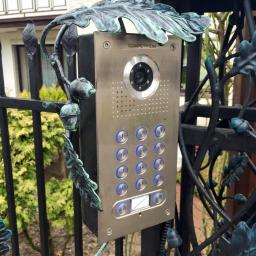 Sarko Krzysztof Okrasa - Panel domofonu z kamerą i klawiaturą numeryczną, zamontowany na metalowym ogrodzeniu z ozdobnymi elementami roślinnymi w kolorze patynowanej miedzi.