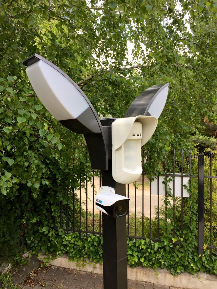 Zestaw oświetleniowo-ochronny na czarnym słupku: kamera BCS, czujnik ruchu i lampa z dwoma białymi reflektorami, na tle zieleni i metalowego ogrodzenia.