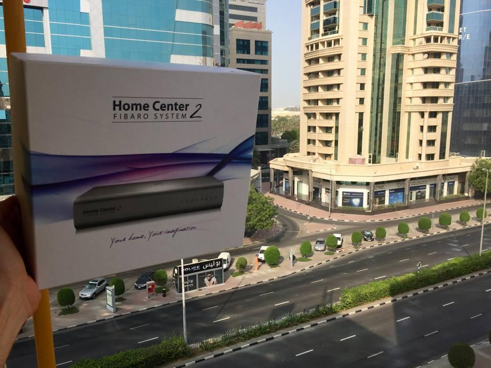 Pudełko z urządzeniem Home Center 2 FIBARO SYSTEM prezentowane na tle miejskiego krajobrazu z wysokimi budynkami i ulicą.