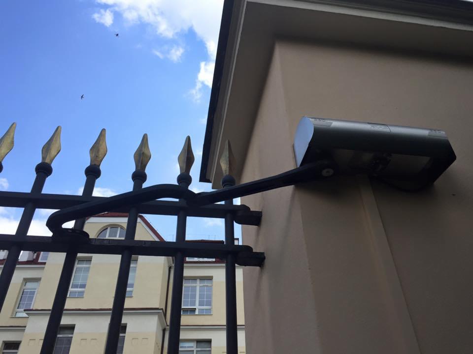 Zewnętrzny reflektor LED zamontowany na elewacji budynku, widoczny przez ozdobne, metalowe ogrodzenie z pozłacanymi grotami.