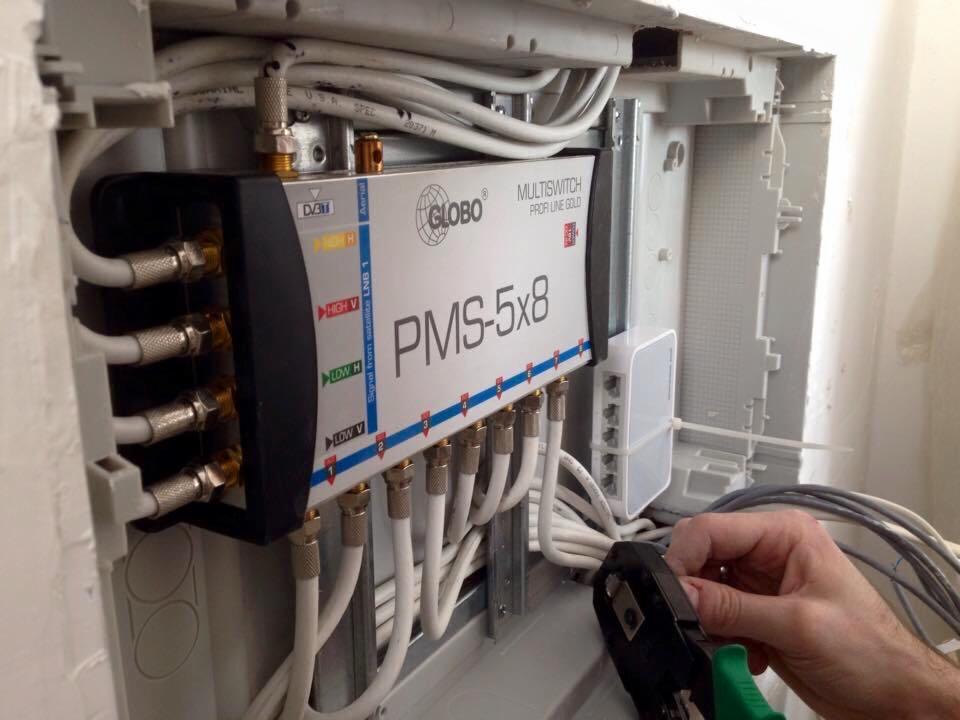 Instalacja multiswitcha Globo PMS-5x8 w skrzynce rozdzielczej, widoczne kable koncentryczne i narzędzie zaciskowe w ręce instalatora.
