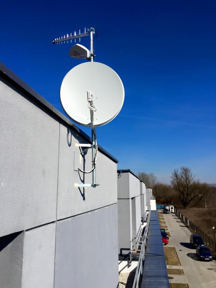 Antena satelitarna i antena kierunkowa zamontowane na szczycie budynku z szarej elewacji, widok z boku, na tle błękitnego nieba.