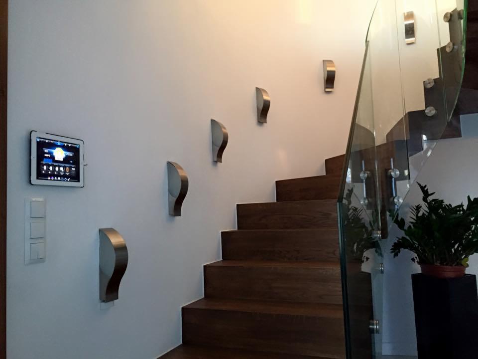 Panel sterowania alarmem domowym, zamontowany na ścianie obok schodów z drewnianymi stopniami i szklaną balustradą, z dodatkowym oświetleniem w postaci metalowych kinkietów.