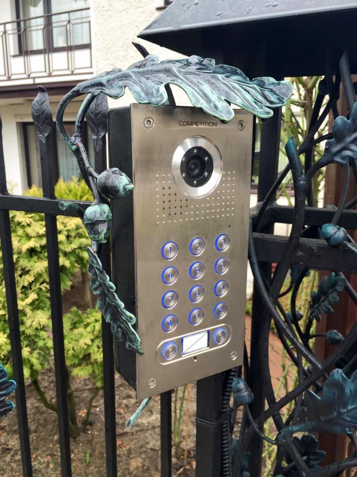 Panel domofonu z kamerą i klawiaturą numeryczną, zamontowany na metalowym ogrodzeniu z ozdobnymi elementami roślinnymi w kolorze patynowanej miedzi.
