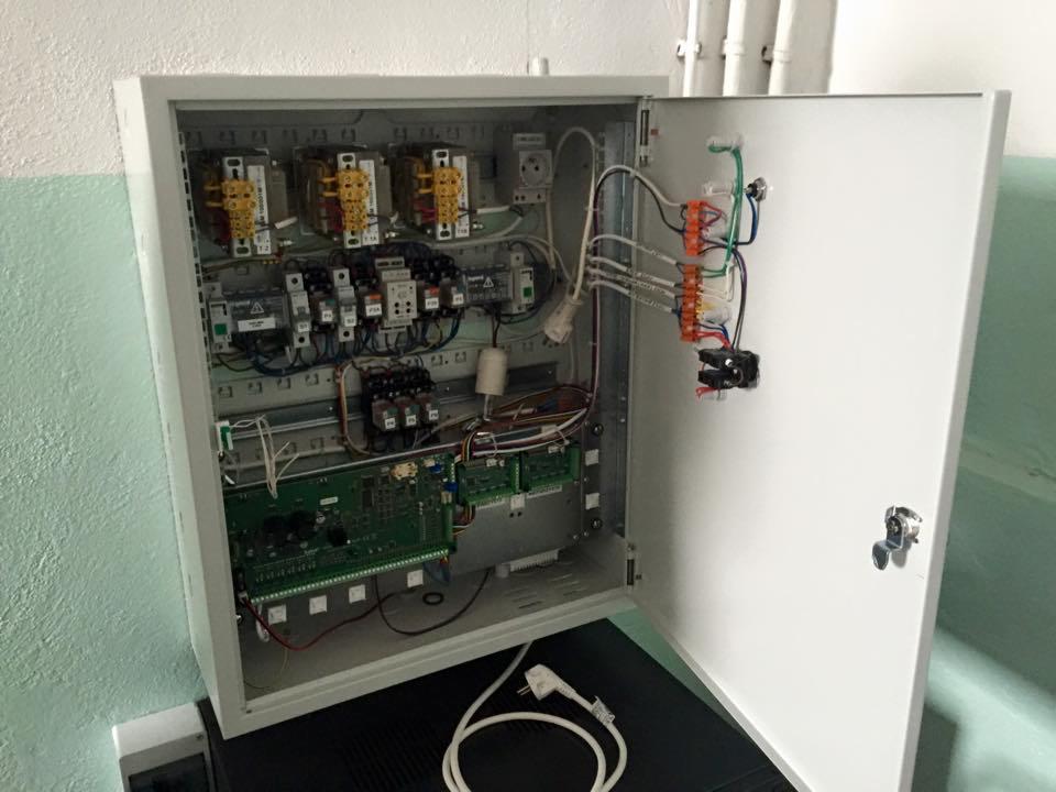 Otwarta skrzynka elektryczna z widocznymi bezpiecznikami, przewodami i płytkami drukowanymi, zamontowana na ścianie z zielonymi akcentami, z przewodem zasilającym na dole.