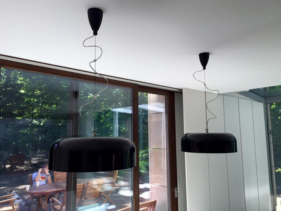 Dwa czarne, nowoczesne klosze lamp wiszących z krętymi przewodami, zamontowane w jasnym wnętrzu z dużymi oknami przesuwnymi wychodzącymi na zielony ogród. W tle widoczna osoba siedząca przy...