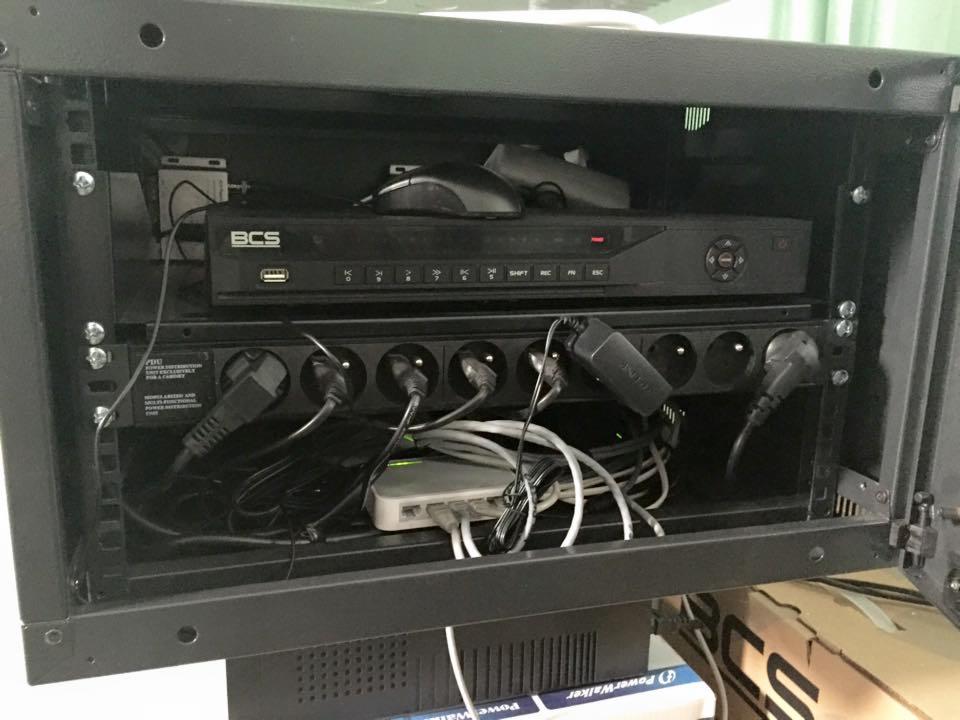 Czarna szafa rack z zamontowanym rejestratorem BCS, listwą zasilającą z podłączonymi wtyczkami, myszką komputerową i routerem, widoczne okablowanie.