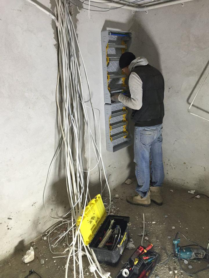 Elektryk w czapce i kamizelce podłącza przewody do rozdzielnicy elektrycznej w surowym pomieszczeniu, z otwartą skrzynką narzędziową i plątaniną kabli na podłodze.