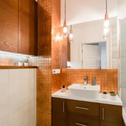 Nowoczesna łazienka z umywalką nablatową, szafką z drewna w ciepłym odcieniu, miedzianą mozaiką na ścianie, wiszącymi lampami z przezroczystego szkła i małymi sukulentami w białych doniczkach.