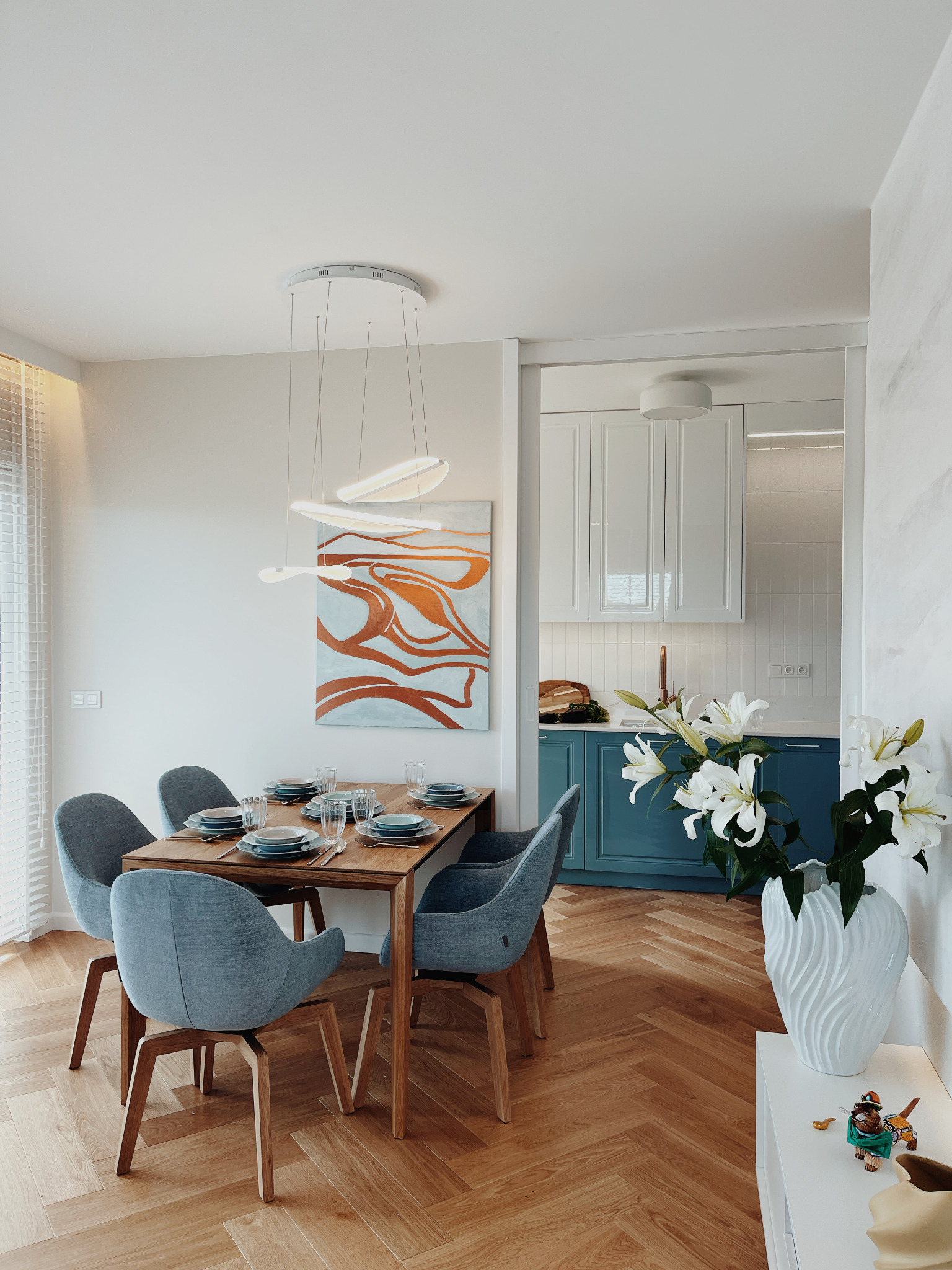 Jadalnia w stylu nowoczesnym: drewniany stół z szarobłękitnymi krzesłami, nad nim designerska lampa, w tle kuchnia z niebieskimi szafkami i białymi liliami.