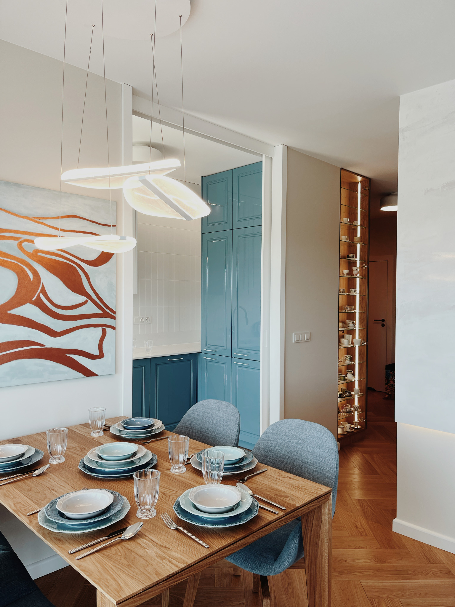 Stół z zastawą w odcieniach błękitu, w tle niebieska zabudowa kuchenna i przeszklona witryna. Całość w jasnym, nowoczesnym wnętrzu z parkietem w jodełkę.