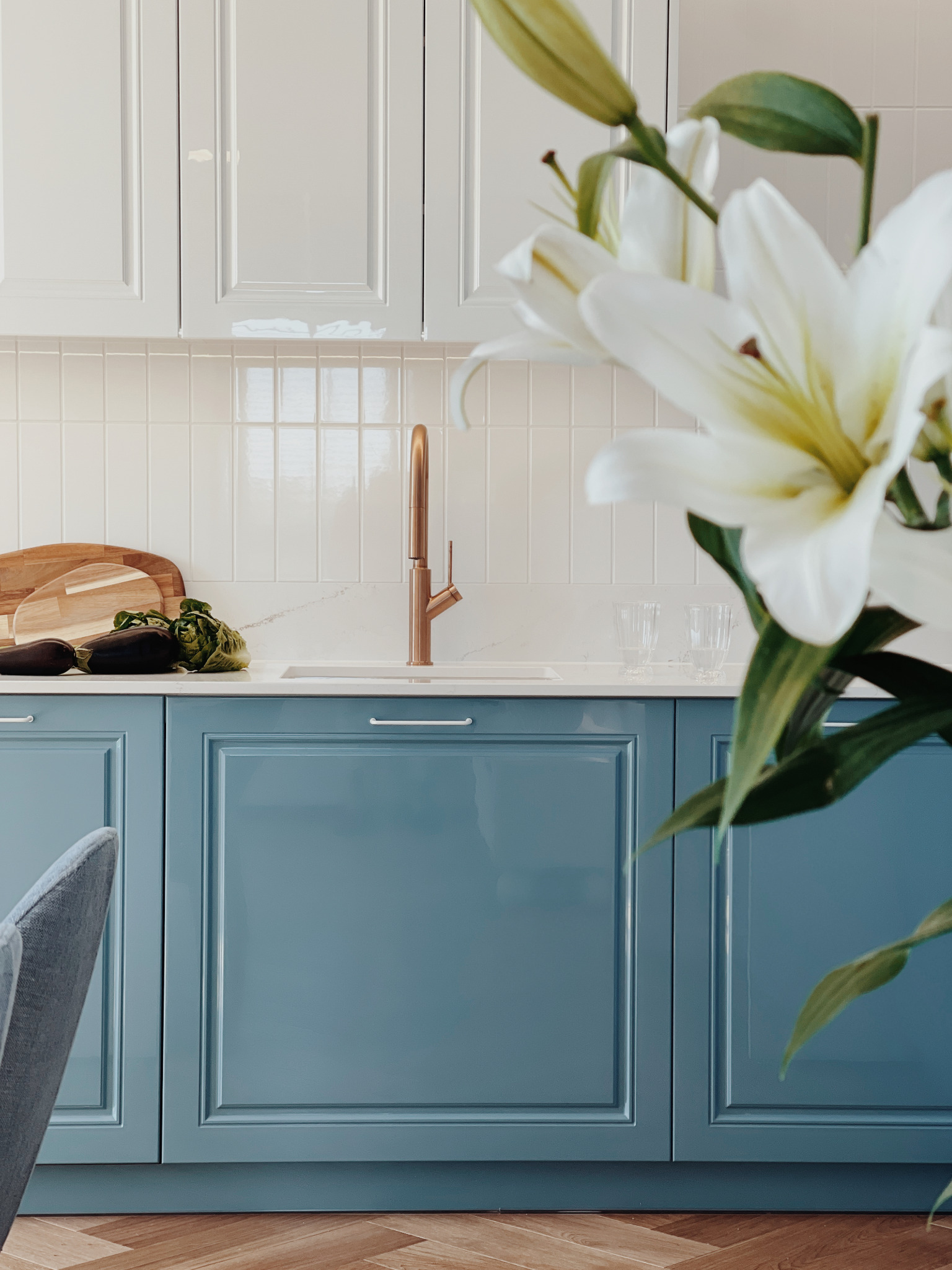 Błękitne szafki kuchenne z miedzianą armaturą, biały blat i płytki. Na blacie warzywa i deski do krojenia. Wazon z białymi liliami w tle. Parkiet w jodełkę.