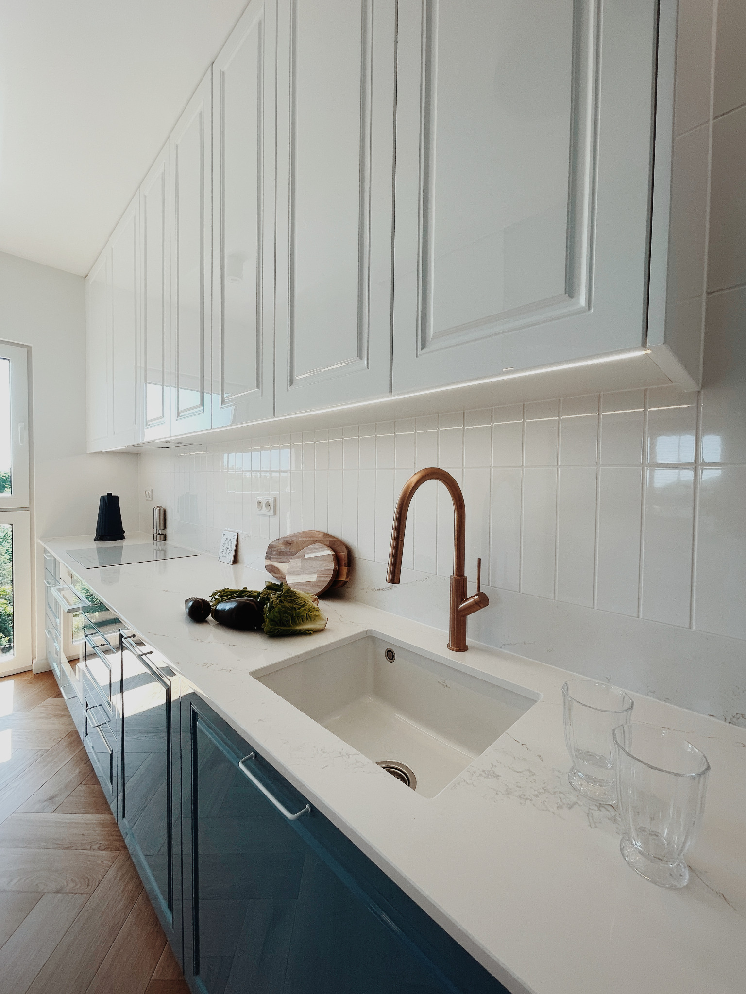 Nowoczesna kuchnia z białymi szafkami, marmurowym blatem i miedzianą armaturą. Na blacie warzywa i szklanki. Parkiet w jodełkę na podłodze.