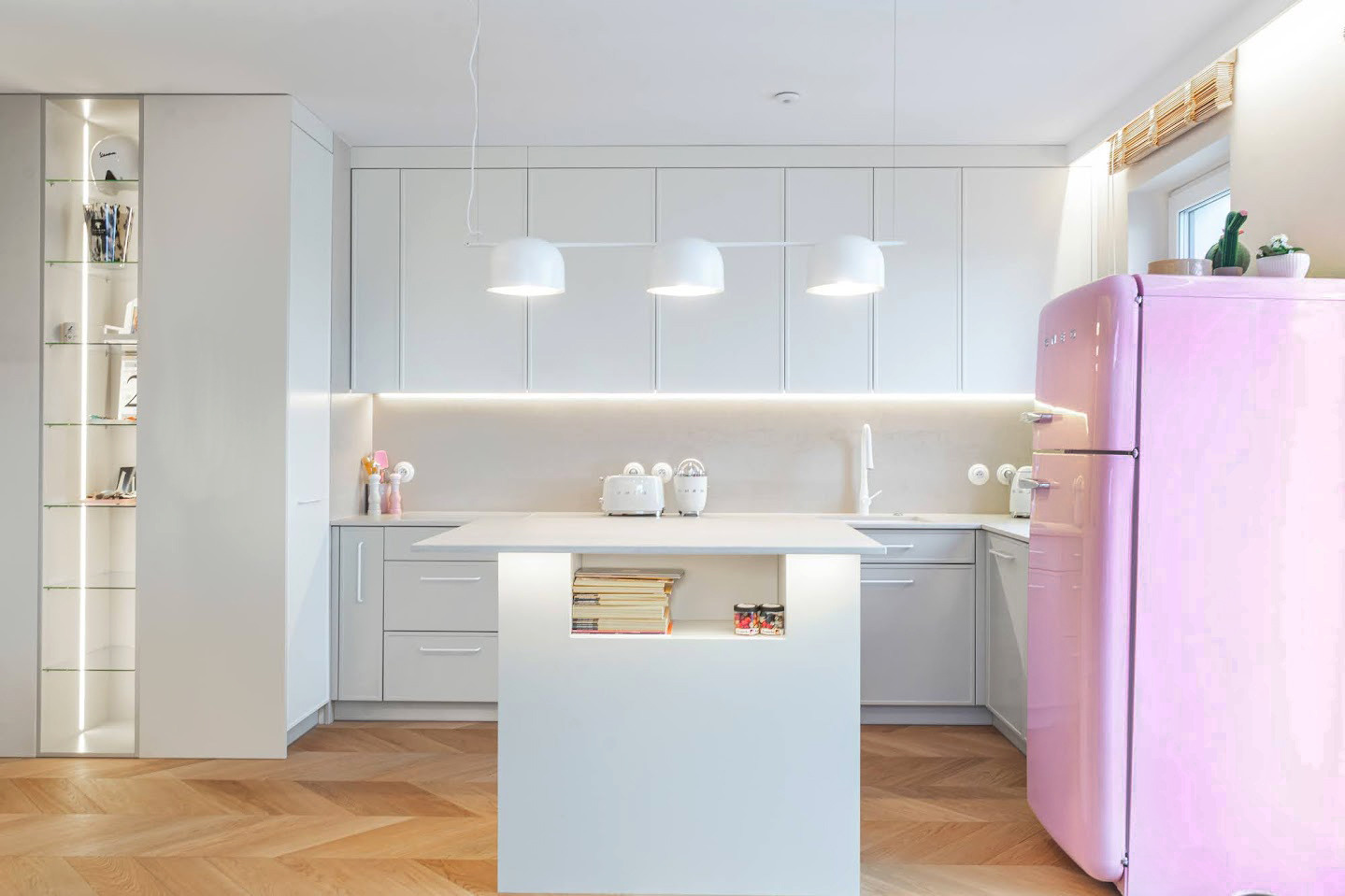 Nowoczesna, jasna kuchnia z wyspą i różową lodówką Smeg. Parkiet w jodełkę, oświetlenie LED pod szafkami, trzy białe lampy wiszące. Minimalistyczny design.