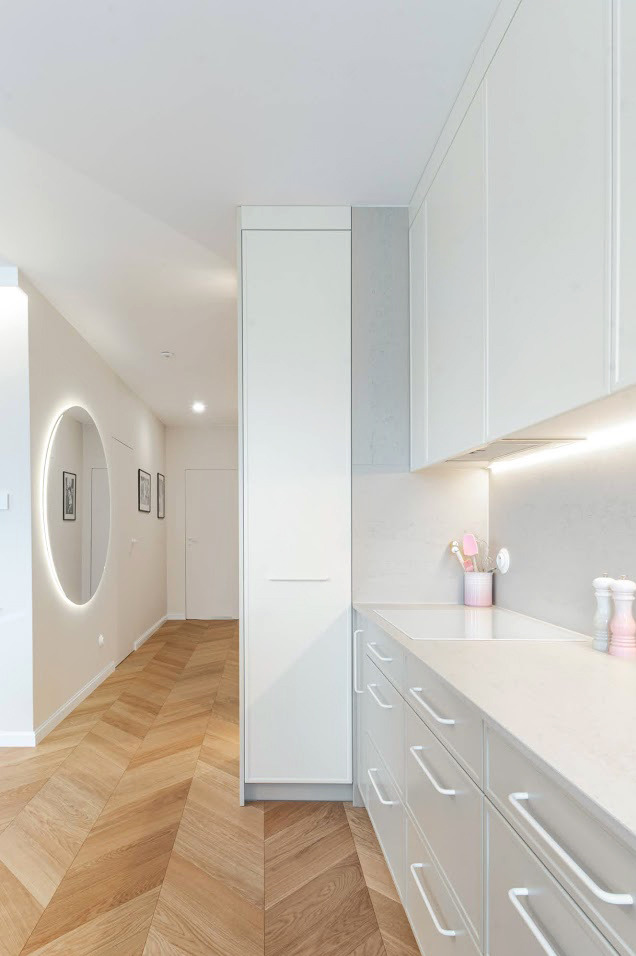 Jasne wnętrze: parkiet w jodełkę, okrągłe lustro z podświetleniem i biała zabudowa kuchenna z delikatnymi, różowymi akcentami. Minimalistyczny design.