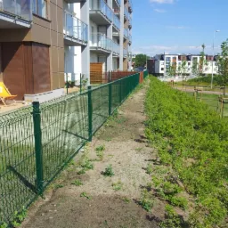 Długi, zielony system ogradzający z siatki, oddzielający budynek mieszkalny od pasa zieleni z młodymi drzewkami.