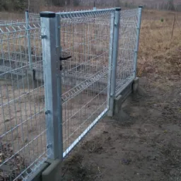 Nowo zamontowana, ocynkowana furtka z paneli kratowych na betonowej podmurówce, widoczna na tle pola i lasu.