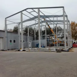 Konstrukcja stalowa hali w trakcie montażu, widoczny żuraw, betonowe słupy i stalowe elementy szkieletu, teren budowy z kontenerem biurowym.