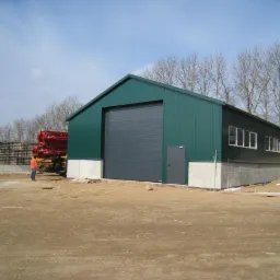 Zielony, blaszany budynek gospodarczy z szarą bramą rolowaną, osadzony na betonowej podmurówce. W tle widoczna betoniarka i osoba w pomarańczowej kurtce.