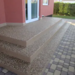 Schody wejściowe z posadzką żywiczną w Gryfinie. Wykończenie z kamyczków, jasne kolory. Widoczny fragment kostki brukowej i elewacji budynku.