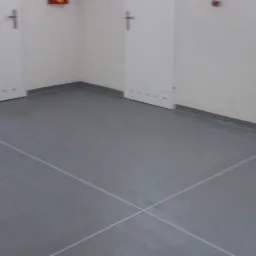 Gładka, szara posadzka z wyraźnymi, białymi liniami krzyżującymi się na środku, przy ścianach białe listwy przypodłogowe i drzwi.