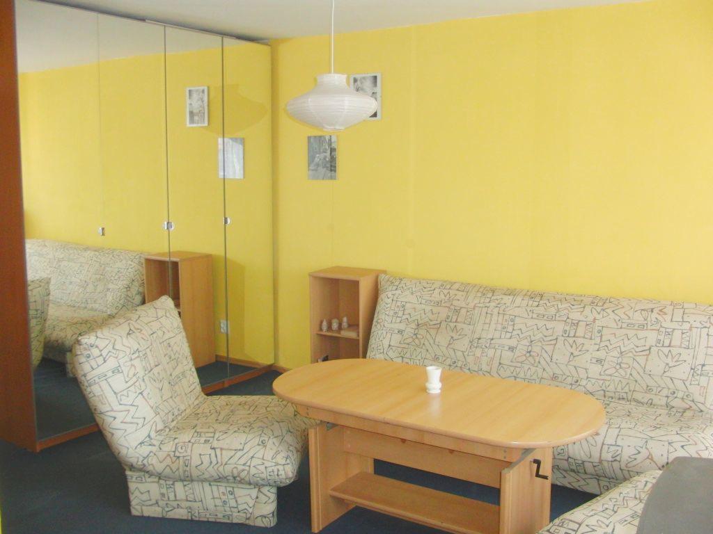 Żółty pokój z wzorzystą sofą i fotelem, drewnianym stołem i szafkami; odbicie sofy w lustrzanej ścianie.